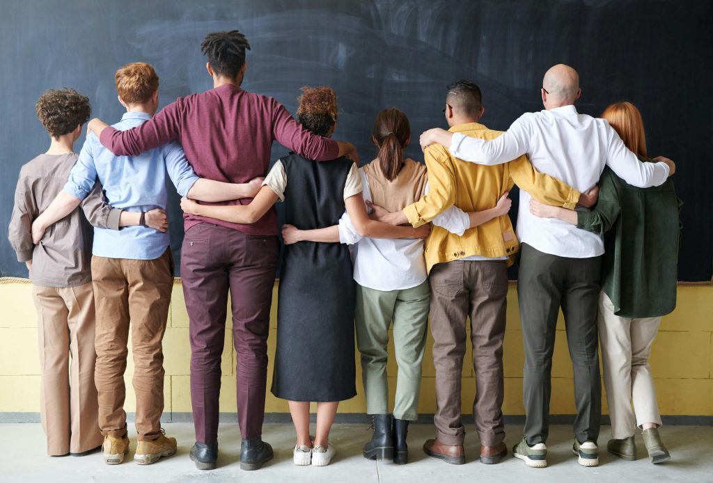 Home A diverse group of adults in casual outfits hugging in front of a chalkboard, symbolizing teamwork.